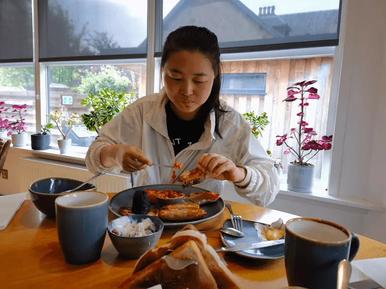 Spring 在窗边餐桌前吃一份含香肠、黑布丁、烤番茄与baked beans 的苏格兰早餐。
