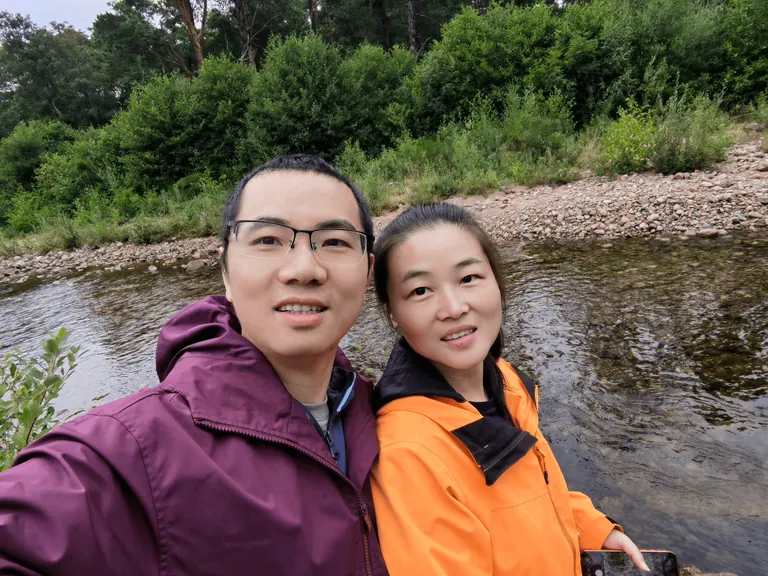 Martin 与 Spring 在浅河弯道前自拍,对岸是卵石滩与密林。