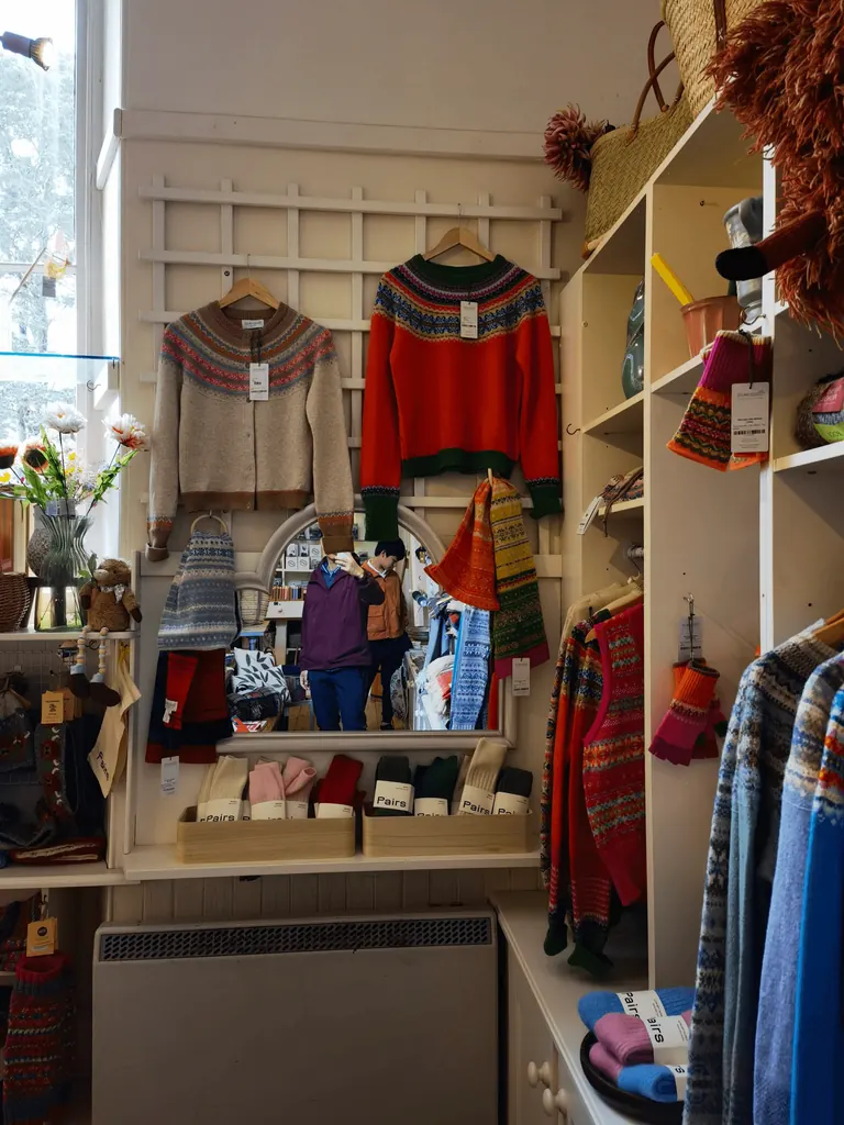针织礼品店内景:Fair Isle 毛衣、成对袜子与白色花格墙架,镜中可见拍摄者。