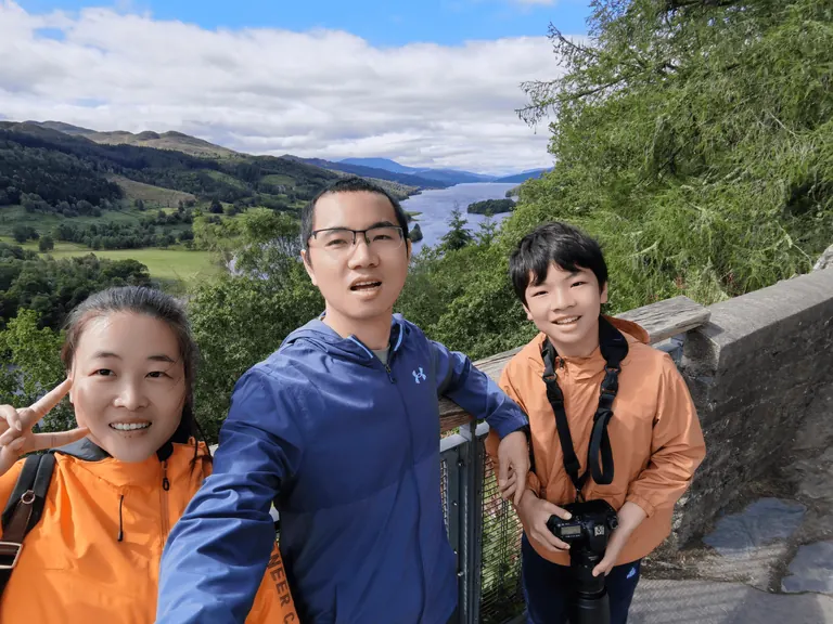 一家三口在 Queen's View 石栏与木质栏杆前自拍,身后是 Loch Tummel 与远山。