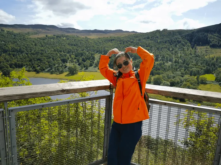 Spring 在 Queen's View 木质栏杆前双手比心,身后是 Loch Tummel 谷地。