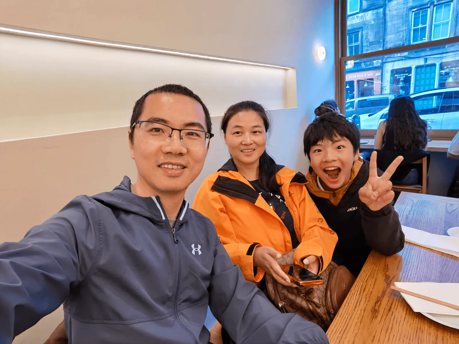 一家三口在居酒屋木桌前自拍,窗外可见 Morrison Street 与 Asian Food & Bar 招牌。