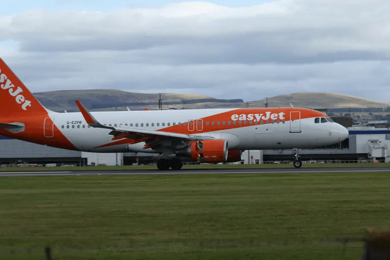 easyJet 客机 G-EZPB 侧影,橙色垂尾,背景是机场建筑与丘陵。